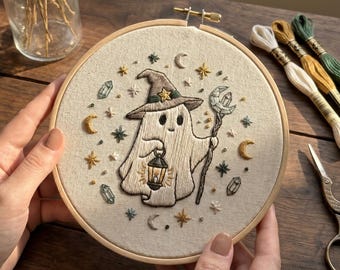 May include: Embroidered ghost wearing a witch hat, holding a lantern and staff, in a wooden embroidery hoop. The design includes stars, moons, and crystals. The artwork is on a beige fabric.