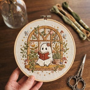 Könnte beinhalten: Gesticktes Bild in einem Holzrahmen, das einen weißen Geist zeigt, der ein Buch in einem Fenster liest, umgeben von Pflanzen und Blumen. Der Geist hat eine Blume auf dem Kopf. Der Hintergrund ist aus Holz. Die Stickerei ist detailliert und farbenfroh.
