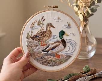 Puede incluir: Un bastidor de bordado circular de madera que contiene un panel de tela con un diseño detallado de dos patos cerca del agua. La escena presenta una pata marrón y un pato real macho con cabeza verde, rodeados de juncos y ondas de agua.