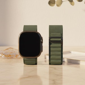 May include: A smartwatch with a dark screen and olive green band is displayed next to its detached band. The watch has a bronze-colored frame. The bands are made of a textured material. The watch and band are on a light-colored surface.