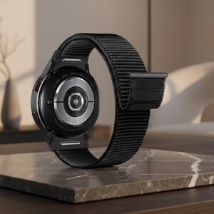 May include: A black smartwatch with a black fabric strap, displayed on a marble surface. The watch face is black with a silver centre. The strap is looped around the watch, highlighting its design. A modern wearable tech accessory.