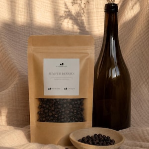 Può includere: Un sacchetto di carta kraft marrone etichettato Soma Herbal Juniper Berries con una finestra trasparente che mostra bacche scure, una bottiglia alta in vetro scuro e una piccola ciotola in ceramica contenente bacche di ginepro essiccate.