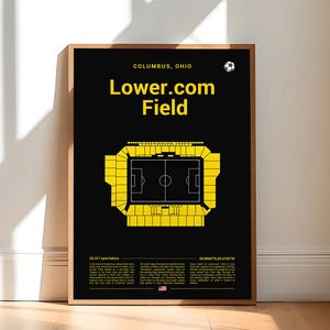 May include: A framed poster with a black background features the text "Lower.com Field" in yellow, above a yellow stadium schematic. The top of the poster reads "Columbus, Ohio" with a soccer ball icon. The poster is leaning against a wall.