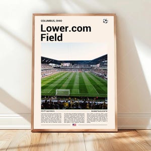 May include: A framed poster featuring Lower.com Field in Columbus, Ohio. The image shows a green soccer field with spectators in the stands. Text at the top reads "COLUMBUS, OHIO" and "Lower.com Field."