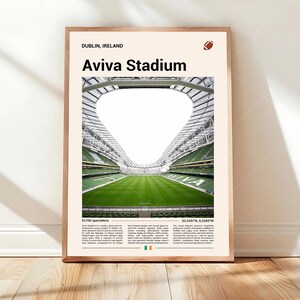 May include: A framed print of Aviva Stadium in Dublin, Ireland. The image features the stadium's green field and seating, with the text "Aviva Stadium" at the top. A football graphic is in the upper right corner. The print is displayed on a wooden floor.