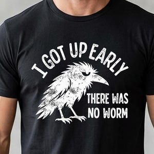 May include: A black short-sleeved t-shirt featuring a white graphic of a grumpy-looking bird. The text printed in white reads, I GOT UP EARLY THERE WAS NO WORM. The design has a distressed, textured appearance on the front of the garment.