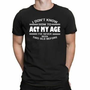 May include: A black short-sleeved t-shirt featuring white text that reads I DON'T KNOW HOW TO ACT MY AGE I'VE NEVER BEEN THIS OLD BEFORE. The design includes horizontal lines above and below the text, centred on the chest of the garment.