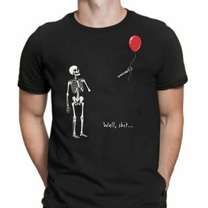 May include: A black short-sleeved t-shirt featuring a graphic of a white skeleton looking at a detached skeletal hand holding a red balloon. The text "Well, shit..." is printed in white below the hand. The design is centred on the chest area.