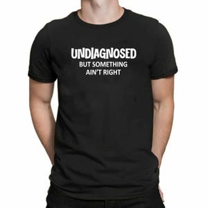 May include: A black short-sleeved t-shirt featuring white text printed on the chest. The text reads UNDIAGNOSED in a large bold font, followed by the smaller text BUT SOMETHING AIN'T RIGHT centred directly underneath the main word.
