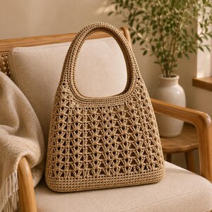 May include: A beige crochet tote bag featuring a structured, open-work fan stitch pattern throughout the body, a solid base, and a single integrated rounded handle. The bag is crafted from a light tan fiber with a consistent, textured weave.