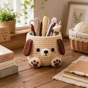 May include: A cream-colored crochet desk organizer shaped like a puppy with dark brown ears, a brown nose, and two front paws with brown paw pads. The puppy face features black eyes and pink cheeks. The container is filled with various pens and scissors.