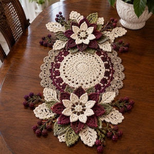 Op de afbeelding: Een gehaakte tafelloper met een centraal rond crèmekleurig kleedje met een geschulpte rand. De loper is aan beide uiteinden versierd met gelaagde driedimensionale bloemen in crème, bordeaux en olijfgroen, geaccentueerd met kleine donkere bessenclusters.
