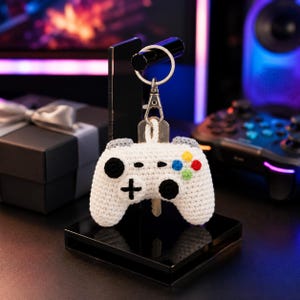 Può includere: Un portachiavi a forma di controller per videogiochi lavorato all'uncinetto in bianco, con pulsanti direzionali neri, un joystick nero e quattro piccoli pulsanti circolari gialli, blu, rossi e verdi. Il controller è fissato a un anello e una chiusura in metallo argentato.