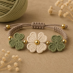 May include: A macrame bracelet featuring a central cream-colored flower motif flanked by two green clover-shaped accents. The design includes small gold-toned metallic beads between the floral elements and a tan adjustable cord band.