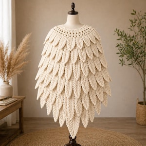 May include: A cream-coloured, handmade crochet cape displayed on a mannequin. The cape has a round neckline and is decorated with multiple rows of leaf-shaped crochet details, creating a textured, layered effect. The cape is displayed on a dark brown mannequin stand.