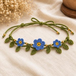 May include: A crochet choker necklace featuring three blue flowers with yellow centers, interspersed with small green crocheted leaves and metallic gold beads along a thin green cord that ties at the back with two long adjustable strings.
