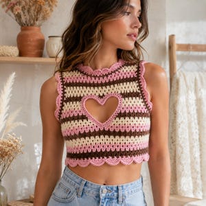 May include: A sleeveless crochet crop top featuring horizontal stripes in shades of pink, cream, and dark brown. The garment has a scalloped edge along the neckline, armholes, and hem, with a prominent heart-shaped cutout centred on the chest.