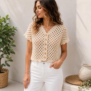 May include: A cream-colored short-sleeve crochet cardigan featuring a V-neckline, a front button closure with six round wooden buttons, and a scalloped edge along the hem and sleeves. The garment is crafted in a classic granny square pattern.