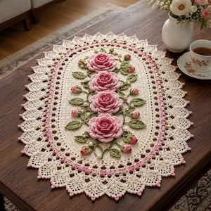 Pode incluir: Um centro de mesa oval em crochê com base na cor creme e borda festonada em rosa e creme. O centro é decorado com quatro rosas tridimensionais, folhas verdes em crochê, pequenos botões de rosa e pérolas brancas espalhadas pelo tecido.