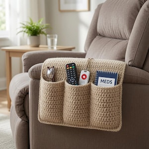 Puede incluir: Un organizador de ganchillo beige con tres bolsillos profundos, colocado sobre el brazo de un sillón reclinable marrón. Los bolsillos contienen unas gafas, un mando a distancia negro, un dispositivo de alerta médica y un cuaderno azul con MEDS.