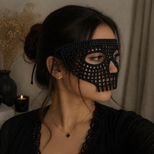 May include: A black crochet skull mask with a detailed open weave design. The mask covers the eyes and nose, with a skeletal jawline. The mask is worn with a black top and a gold necklace.