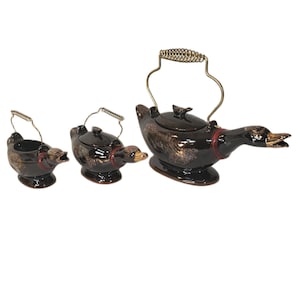 May include: A set of three ceramic duck-shaped teapots and a creamer. The teapots are dark brown with gold accents and a red band around the neck. The largest teapot has a gold handle, and the smallest is a creamer.