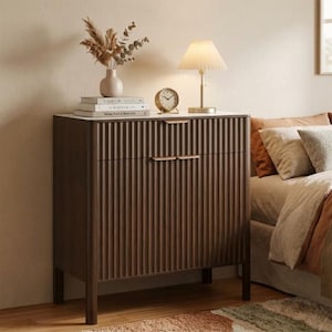 Puede incluir: Una mesita de noche de madera marrón oscuro con tapa blanca, frente acanalado y dos cajones. Un pequeño reloj dorado, una lámpara con pantalla plisada y un jarrón con flores secas encima. La mesita de noche está en un dormitorio.