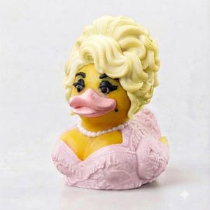 Könnte beinhalten: Eine gelbe Quietscheente mit einer voluminösen blonden Perücke, dunklen Wimpern und einem rosa Schnabel. Die Ente trägt eine weiße Perlenkette und ein strukturiertes, hellrosa Kleid mit spitzenartigen Details am Oberteil und an den Flügeln.