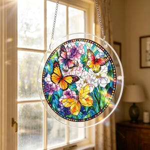 May include: A round, stained-glass window hanging featuring colorful butterflies and flowers. The butterflies are yellow, orange, and pink, set against a backdrop of purple, blue, and white flowers. The window is suspended by a silver chain.