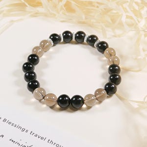 May include: A beaded bracelet featuring alternating black and smoky quartz beads. The bracelet has silver-colored spacers and is displayed on a white surface. The beads are round and polished, creating a contrast in color and texture.