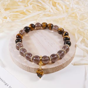 Könnte beinhalten: Ein Armband mit braunen, grauen und Tigeraugen-Perlen. Es hat einen goldfarbenen Anhänger mit einem Tigeraugen-Anhänger. Die Perlen sind durch goldfarbene Abstandshalter getrennt.