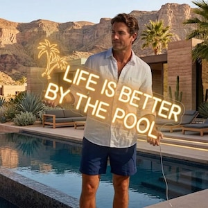 以下が含まれることがあります： 「LIFE IS BETTER BY THE POOL」の文字が筆記体で書かれたネオンサイン。ヤシの木のグラフィック付き。プールと山の背景を前に、人がそのサインを持っています。