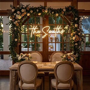 May include: A wooden dining table with two upholstered chairs featuring Mr. and Mrs. labels, set beneath a large floral archway adorned with greenery and roses. A glowing neon sign displays the text The Smiths in a cursive script font.