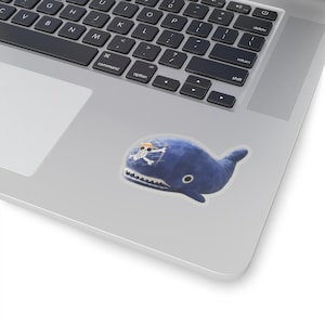 May include: A blue plush whale sticker with a white skull and crossbones design wearing a straw hat. The whale has a white mouth and a black eye. The sticker is on a silver laptop.
