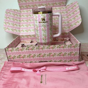 Puede incluir: Un termo de viaje Stanley LoveShackFancy rosa con estampado floral y asa, dentro de una caja a juego. La caja y el termo presentan un patr&oacute;n floral rosa y blanco. Tambi&eacute;n se incluye una correa rosa con los logotipos de Stanley y LoveShackFancy.