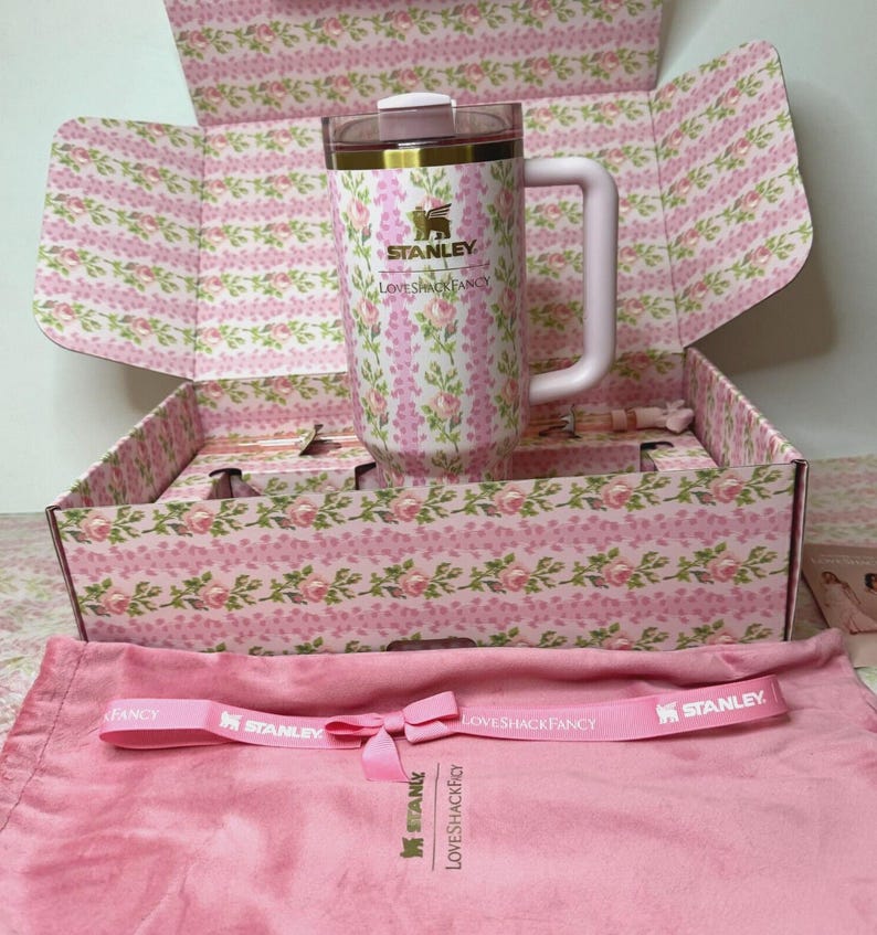 Puede incluir: Un termo de viaje Stanley LoveShackFancy rosa con estampado floral y asa, dentro de una caja a juego. La caja y el termo presentan un patr&oacute;n floral rosa y blanco. Tambi&eacute;n se incluye una correa rosa con los logotipos de Stanley y LoveShackFancy.