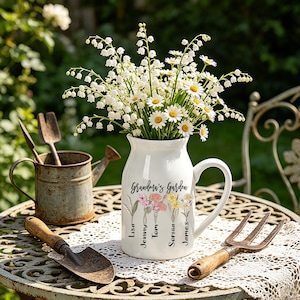 May include: A white ceramic jug with a handle, filled with white and yellow flowers, and the words "Grandma's Garden". The jug has floral illustrations and names. A small trowel, a gardening fork, and a watering can are nearby.