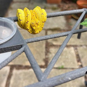 May include: A bright yellow snake with small dark brown markings coiled tightly around a thin grey metal bar. The snake has a triangular head and a slender body, resting securely on the textured surface of the grey metal structure.