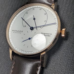Pu&ograve; includere: Un orologio da polso Nomos Glash&uuml;tte con cassa color oro rosa, quadrante bianco argento e cinturino in pelle marrone scuro. Il quadrante presenta sottili lancette blu scuro, un indicatore della riserva di carica e piccoli indici neri.
