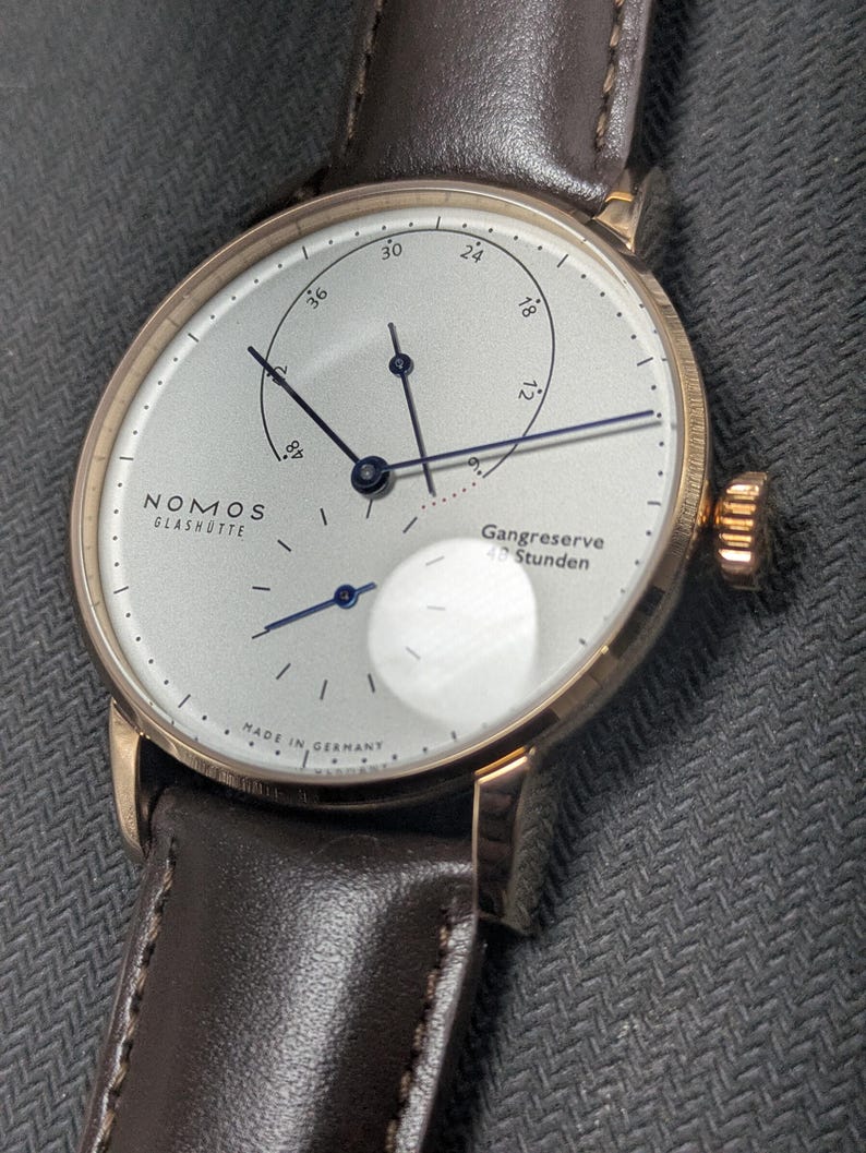 Pu&ograve; includere: Un orologio da polso Nomos Glash&uuml;tte con cassa color oro rosa, quadrante bianco argento e cinturino in pelle marrone scuro. Il quadrante presenta sottili lancette blu scuro, un indicatore della riserva di carica e piccoli indici neri.