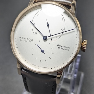 Pu&ograve; includere: Un orologio da polso Nomos Glash&uuml;tte con cassa rotonda color oro, quadrante bianco e cinturino in pelle marrone scuro. Il quadrante presenta sottili indici neri, lancette blu, un indicatore della riserva di carica con il testo Gangreserve 48 Stunden e Made in Germany.
