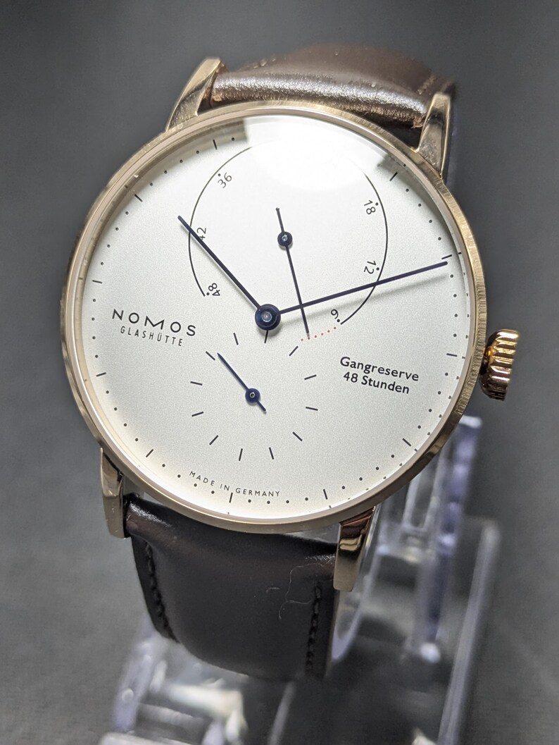 Pu&ograve; includere: Un orologio da polso Nomos Glash&uuml;tte con cassa rotonda color oro, quadrante bianco e cinturino in pelle marrone scuro. Il quadrante presenta sottili indici neri, lancette blu, un indicatore della riserva di carica con il testo Gangreserve 48 Stunden e Made in Germany.