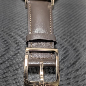 Pu&ograve; includere: Un orologio da polso con quadrante bianco e cassa color oro rosa. L'orologio presenta un cinturino in pelle color tortora scuro con cuciture tono su tono e una fibbia lucida color oro rosa. La parola NOMOS &egrave; incisa sulla fibbia metallica.