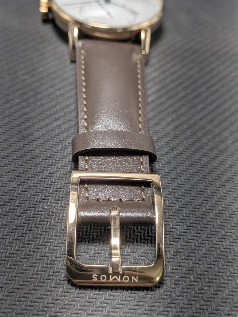 Pu&ograve; includere: Un orologio da polso con quadrante bianco e cassa color oro rosa. L'orologio presenta un cinturino in pelle color tortora scuro con cuciture tono su tono e una fibbia lucida color oro rosa. La parola NOMOS &egrave; incisa sulla fibbia metallica.