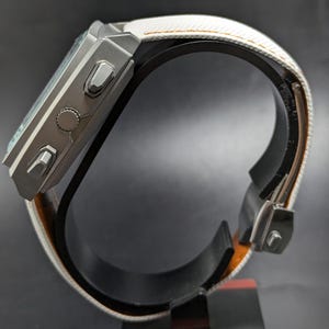 May include: A digital wristwatch with a matte grey rectangular case featuring three side buttons and a textured white strap with orange stitching. The watch is displayed from a side profile view, showing the metallic casing and the strap buckle mechanism.