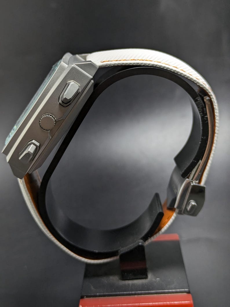 May include: A digital wristwatch with a matte grey rectangular case featuring three side buttons and a textured white strap with orange stitching. The watch is displayed from a side profile view, showing the metallic casing and the strap buckle mechanism.