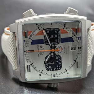 May include: A square-cased TAG Heuer Monaco chronograph watch with a silver dial, featuring blue and orange racing stripes, two black sub-dials, a date window at the three o'clock position, and a white textured strap with orange stitching.