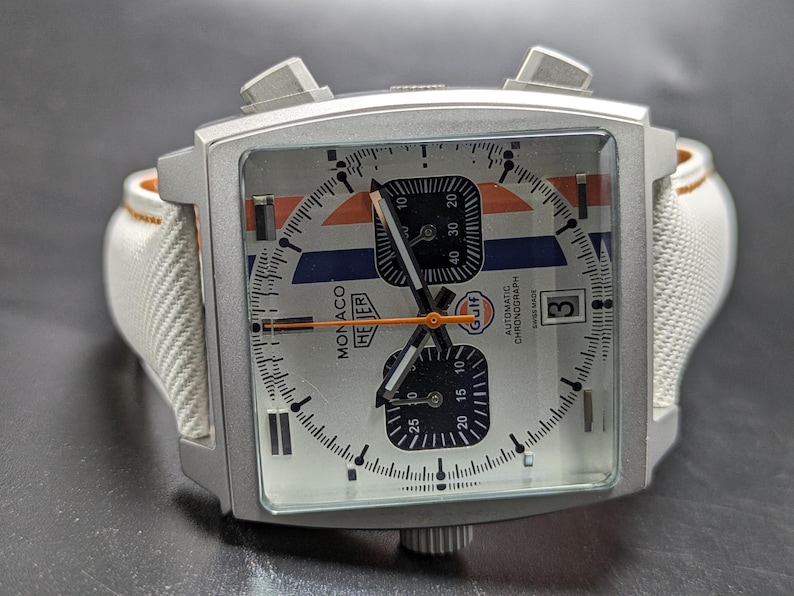 May include: A square-cased TAG Heuer Monaco chronograph watch with a silver dial, featuring blue and orange racing stripes, two black sub-dials, a date window at the three o'clock position, and a white textured strap with orange stitching.