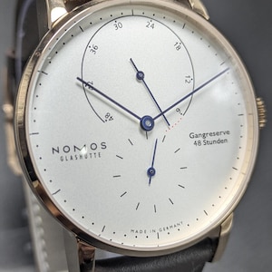 Pu&ograve; includere: Un orologio da polso Nomos Glash&uuml;tte con cassa rotonda color oro, quadrante bianco e cinturino in pelle marrone scuro. Il quadrante presenta lancette blu, un indicatore della riserva di carica con il testo Gangreserve 48 Stunden e Made in Germany.