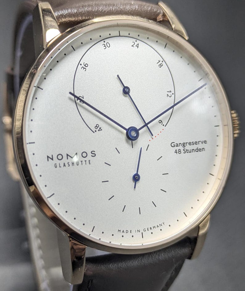 Pu&ograve; includere: Un orologio da polso Nomos Glash&uuml;tte con cassa rotonda color oro, quadrante bianco e cinturino in pelle marrone scuro. Il quadrante presenta lancette blu, un indicatore della riserva di carica con il testo Gangreserve 48 Stunden e Made in Germany.